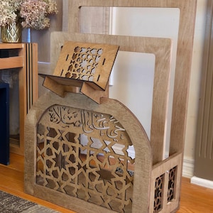 May include: A wooden book stand with intricate laser cut designs. The stand has a shelf for a book and a decorative arch with Arabic calligraphy. The stand is made of light brown wood.