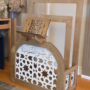 May include: A wooden Quran stand with intricate laser cut designs. The stand features a shelf for a book and a decorative arch with Arabic calligraphy. The stand is made of light brown wood and has a white background.