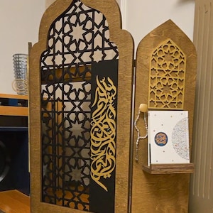 Peut inclure: Un séparateur de pièce en bois à trois panneaux avec des motifs géométriques découpés au laser complexes en or et noir. Le panneau central présente une calligraphie arabe en or. Une petite étagère sur le panneau de droite contient un livre et des chapelets.