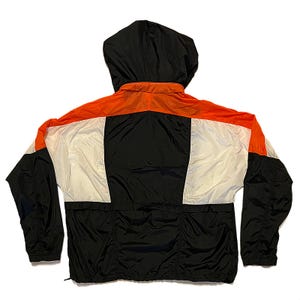 K&ouml;nnte beinhalten: Windjacke mit Kapuze in Schwarz, Wei&szlig; und Orange. Die R&uuml;ckseite der Jacke ist sichtbar und zeigt ein farbblockiertes Design. Die Kapuze ist schwarz, und die Jacke hat einen durchgehenden Rei&szlig;verschluss und elastische B&uuml;ndchen.