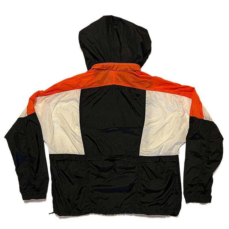 K&ouml;nnte beinhalten: Windjacke mit Kapuze in Schwarz, Wei&szlig; und Orange. Die R&uuml;ckseite der Jacke ist sichtbar und zeigt ein farbblockiertes Design. Die Kapuze ist schwarz, und die Jacke hat einen durchgehenden Rei&szlig;verschluss und elastische B&uuml;ndchen.