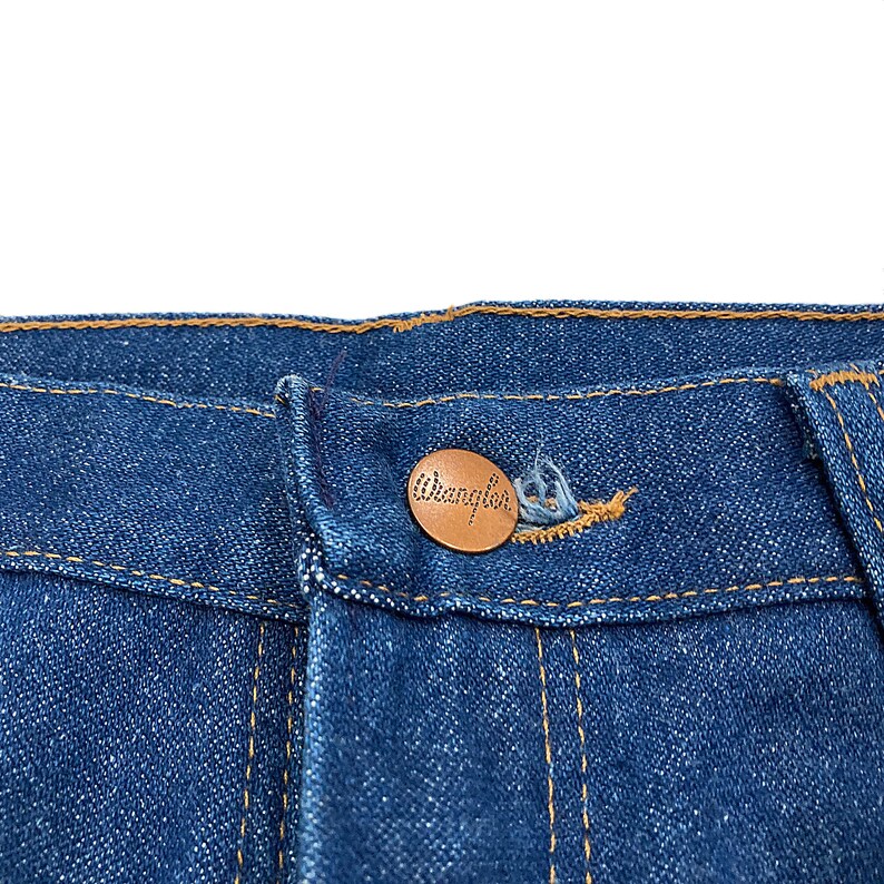 K&ouml;nnte beinhalten: Nahaufnahme einer blauen Jeans mit einem kupferfarbenen Knopf. Der Knopf ist mit dem Markennamen "Wrangler" gepr&auml;gt. Die Jeans hat gelbe N&auml;hte und ein klassisches Design.