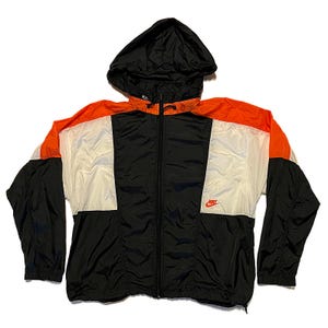 K&ouml;nnte beinhalten: Eine schwarze, orange und wei&szlig;e Nike Windjacke mit Kapuze. Die Jacke hat einen durchgehenden Rei&szlig;verschluss und das rote Nike-Logo auf der Brust. Die Jacke ist aus leichtem Material, m&ouml;glicherweise Nylon.