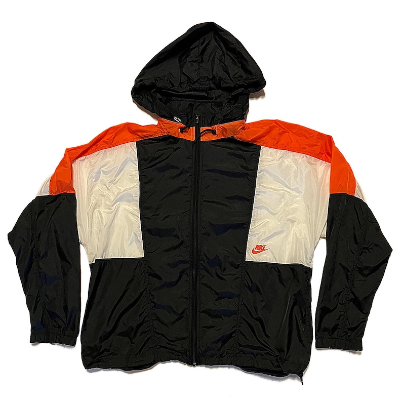 K&ouml;nnte beinhalten: Eine schwarze, orange und wei&szlig;e Nike Windjacke mit Kapuze. Die Jacke hat einen durchgehenden Rei&szlig;verschluss und das rote Nike-Logo auf der Brust. Die Jacke ist aus leichtem Material, m&ouml;glicherweise Nylon.