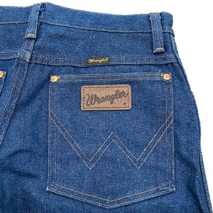 K&ouml;nnte beinhalten: Nahaufnahme der R&uuml;ckseite einer dunkelblauen Jeans. Die Jeans hat einen braunen Lederaufn&auml;her mit dem Markennamen "Wrangler" und ein kleines schwarzes Etikett mit dem Markennamen &uuml;ber der Ges&auml;&szlig;tasche. Die N&auml;hte sind in einer kontrastierenden beigen Farbe gehalten.