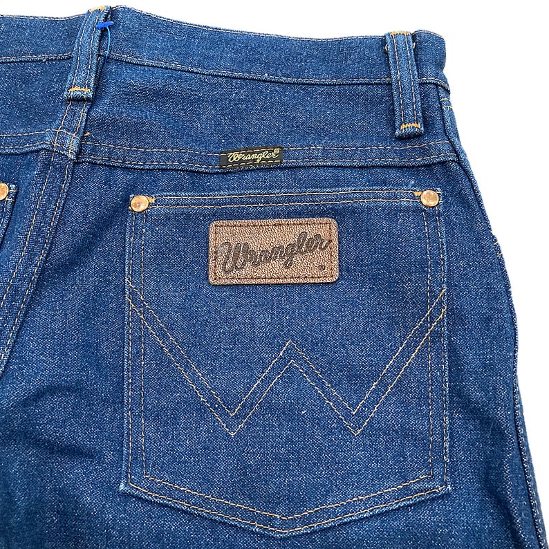 K&ouml;nnte beinhalten: Nahaufnahme der R&uuml;ckseite einer dunkelblauen Jeans. Die Jeans hat einen braunen Lederaufn&auml;her mit dem Markennamen "Wrangler" und ein kleines schwarzes Etikett mit dem Markennamen &uuml;ber der Ges&auml;&szlig;tasche. Die N&auml;hte sind in einer kontrastierenden beigen Farbe gehalten.