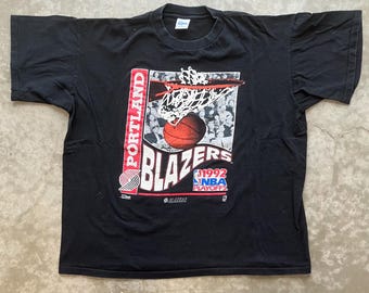 Puede incluir: Camiseta negra con un gráfico de baloncesto de los Portland Blazers. El gráfico incluye una pelota de baloncesto entrando en una red, la palabra "BLAZERS" en blanco y "1992 NBA Playoffs". La palabra "PORTLAND" está impresa verticalmente.