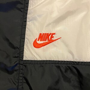 K&ouml;nnte beinhalten: Nahaufnahme einer schwarzen, wei&szlig;en und schwarzen Sportjacke. Das Nike-Logo ist orange auf dem wei&szlig;en Feld gestickt. Die Jacke hat eine Nahtdetail.