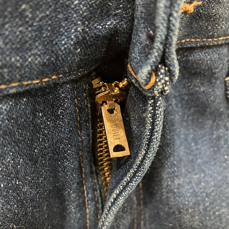 K&ouml;nnte beinhalten: Nahaufnahme einer blauen Jeans mit einem Messingrei&szlig;verschluss. Der Rei&szlig;verschlussanh&auml;nger tr&auml;gt die Marke "SCOVILL". Der Jeansstoff weist Gebrauchsspuren und Ausbleichungen auf.