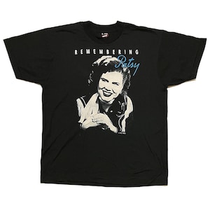 May include: Black t-shirt featuring a black and white image of a woman with the text "REMEMBERING Patsy" in white and blue. The image is a portrait of a smiling woman. The t-shirt is made of a soft material.