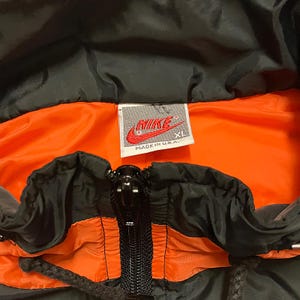 K&ouml;nnte beinhalten: Nahaufnahme einer schwarzen und orangefarbenen Nike Jacke. Die Innenseite ist leuchtend orange, w&auml;hrend die Au&szlig;enseite schwarz ist. Ein Nike-Etikett ist sichtbar, mit dem Text "MADE IN U.S.A." und der Gr&ouml;&szlig;e "XL". Die Jacke hat einen durchgehenden Rei&szlig;verschluss.