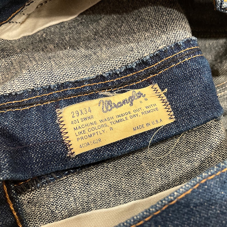 K&ouml;nnte beinhalten: Nahaufnahme einer blauen Jeans. Das Etikett zeigt die Marke "Wrangler", die Gr&ouml;&szlig;e 29x34 Zoll und Waschanweisungen. Die Jeans ist in den USA hergestellt.