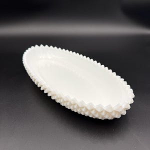 May include: A white, oval-shaped dish with a textured, sawtooth-like edge. The dish is made of a solid, opaque material and sits on a dark surface. The dish is empty and appears to be a decorative serving piece.