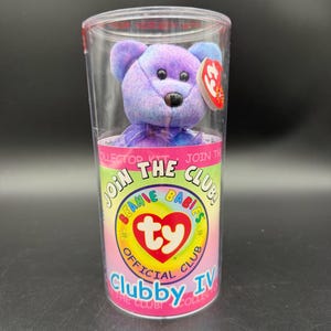May include: A purple tie-dye plush bear with black eyes and nose, wearing a red heart-shaped tag, enclosed in a clear plastic cylindrical container labeled Join The Club Beanie Babies Official Club Clubby IV with a rainbow graphic design.