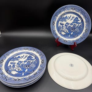 May include: A collection of blue and white ceramic plates with a classic Willow pattern design. The plates feature a central scene with pagodas, trees, and birds, surrounded by a decorative border. One plate is on a red stand.
