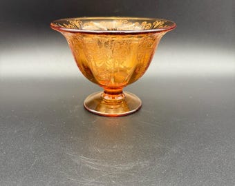 Pode incluir: Uma tigela de vidro âmbar, com pé e um padrão floral decorativo. A tigela tem uma borda larga e alargada e uma base redonda. O vidro é transparente, permitindo a passagem da luz, e está sobre um fundo escuro.