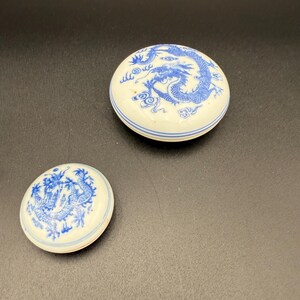 May include: Two white ceramic trinket boxes with blue dragon designs. The larger box has a detailed dragon illustration on the lid, while the smaller box features a similar design. Both boxes have a blue stripe around the edge.