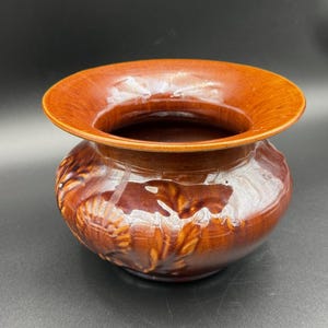 May include: A brown ceramic planter with a wide, flared rim and a rounded body. The planter has a glossy finish and a textured, embossed design around the base. The color is a warm, earthy tone.
