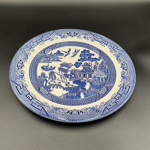 May include: A round, blue and white ceramic plate with a Willow pattern design. The plate features a central scene of a pagoda, bridge, and figures, surrounded by a decorative border. The plate is set against a dark background.