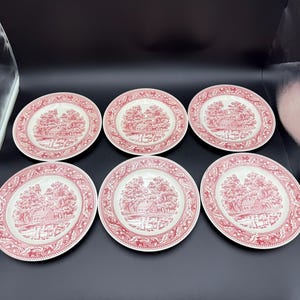 May include: A set of six round ceramic plates featuring a red and white transferware pattern. Each plate displays a central landscape illustration of a house surrounded by trees, framed by a decorative border of floral and animal motifs.