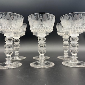 May include: A set of six clear crystal stemmed glasses. Each glass features a floral design etched into the bowl, a twisted stem, and a faceted base. The glasses are arranged on a dark surface.