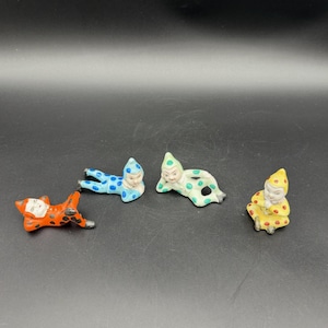 May include: Four small ceramic figurines of jesters in different poses and colors. One is orange with black spots, one is blue with blue spots, one is light green with green spots, and one is yellow with red spots. Each wears a pointed hat.
