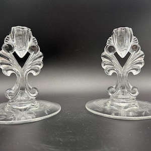May include: Two clear glass candle holders with ornate, stylized leaf and scroll designs. The candle holders have a wide, flat base with etched floral patterns. The candle holders are made of clear glass and are on a dark background.