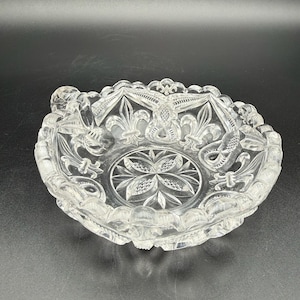 May include: Clear glass ashtray with a detailed fleur-de-lis and floral design. The ashtray has a scalloped edge and a central starburst pattern. The clear glass allows light to pass through, highlighting the intricate carvings.