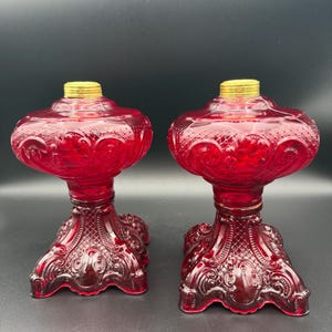 May include: Two antique ruby red glass oil lamps. Each lamp features a decorative, ornate base and a bulbous reservoir. The lamps have a gold-colored metal top. The lamps are displayed against a dark background.