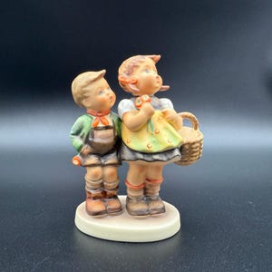 May include: A ceramic figurine of two children, a boy and a girl, standing side by side. The boy wears a green jacket, brown overalls, and boots. The girl wears a yellow dress and holds a basket. The figurine is on a white base.