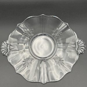 May include: Clear glass serving dish with a scalloped edge and decorative leaf accents. The dish has a central recessed area and two handles. The clear glass allows for visibility of the contents.