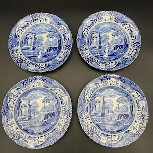 May include: Four blue and white decorative plates. Each plate features a detailed landscape scene with architectural elements and figures. The plates have a floral border design and are arranged on a dark surface.