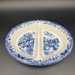 May include: An oval ceramic divided serving dish featuring a cream-colored base with blue transferware patterns. The rim displays a floral and wheat motif, while the two interior sections depict detailed rural landscape scenes with figures.