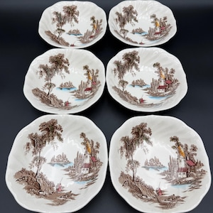 May include: Six white, scalloped-edge bowls with a brown and sepia landscape design. The design features trees, buildings, and figures near a body of water. The bowls are arranged on a dark surface.