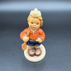 May include: A ceramic figurine of a young boy wearing a white hat, orange shirt, and blue shorts. The boy is holding a wooden shovel. The figurine is standing on a white circular base.