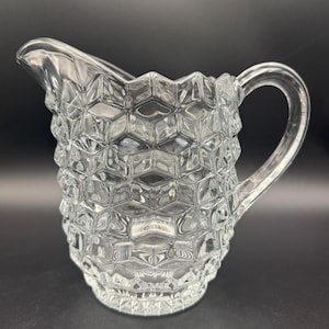 May include: A clear glass pitcher with a honeycomb pattern. The pitcher has a spout and a curved handle. The glass is transparent, allowing light to pass through. The pitcher is likely used for serving beverages.