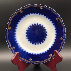 May include: A vintage porcelain plate with a scalloped edge, featuring a deep blue border and a white center. The blue section has a detailed pattern, and the rim is accented with gold trim. The plate is displayed on a red wooden stand.