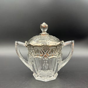 May include: A clear glass sugar bowl with a matching lid, featuring two side handles and a faceted finial. The upper portion of the bowl and the lid are decorated with metallic silver trim in a geometric and floral pattern, with vertical fluting.
