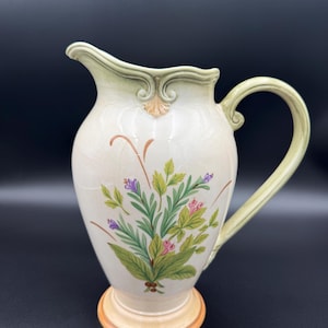May include: Cream-colored ceramic pitcher with a floral design. The pitcher features a light green handle and rim, and a base with a tan and orange trim. The floral design includes green leaves, purple and pink flowers, and brown stems.