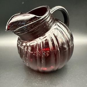 May include: A ruby red glass pitcher with a rounded body and a ribbed design. The pitcher has a curved handle and a spout for pouring. The glass is transparent, allowing light to pass through. The pitcher is likely used for serving beverages.