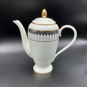 May include: White porcelain coffee pot with a gold finial and trim. The pot features a black and gold geometric pattern around the middle. The handle and spout are white. The pot sits on a small pedestal base.
