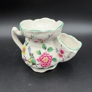 May include: A white ceramic shaving scuttle with a floral and bird design. The scuttle has a handle and two compartments, one for soap and one for water. The floral design includes pink and yellow flowers with green leaves.