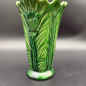 May include: A vibrant green ceramic vase featuring a raised, molded botanical pattern of leaves and flower buds. The vase has a scalloped, wavy rim and a circular base, with a glossy, reflective glaze that highlights the intricate relief texture.