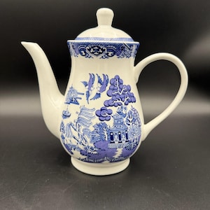 May include: White ceramic teapot with a blue and white Willow pattern design. The teapot features a curved spout, a rounded handle, and a lid with a small knob. The design includes scenes of pagodas, trees, and birds.