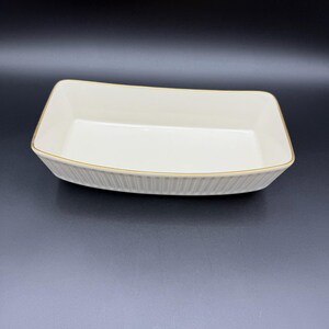 May include: Cream-colored rectangular serving dish with a gold trim. The dish has a slightly curved shape and vertical ribbed detailing on the sides. The interior is smooth and the overall design is simple and elegant.