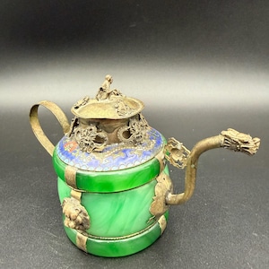 May include: An antique teapot with a green jade-like body and ornate metal accents. The lid and spout feature intricate dragon and floral designs. The handle is a curved metal piece. The teapot has a blue and gold rim.