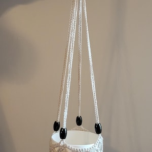 May include: A white macrame plant hanger with a wooden ring at the top and black wooden beads. The hanger is designed to hold a small pot.