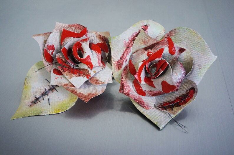May include: Two white fabric roses with red paint splatters and stitched details. The roses have a distressed, vintage look.