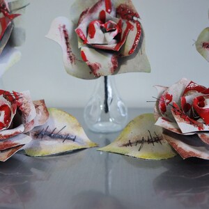 May include: Five white roses with red paint and stitched details are arranged in clear glass vases. The roses have a distressed, vintage look.