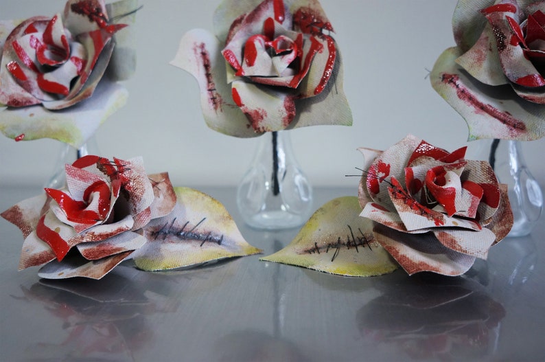 May include: Five white roses with red paint and stitched details are arranged in clear glass vases. The roses have a distressed, vintage look.